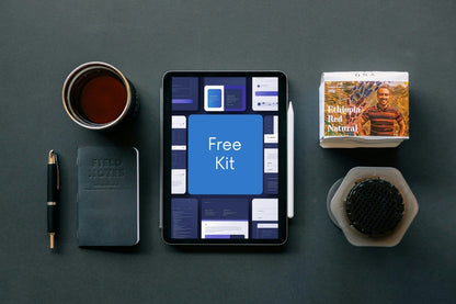 A styled flat-lay featuring a tablet displaying a digital design template kit labeled 'Free Kit', surrounded by a black notebook, a pen, a cup of tea, and a small book on a dark gray surface.
